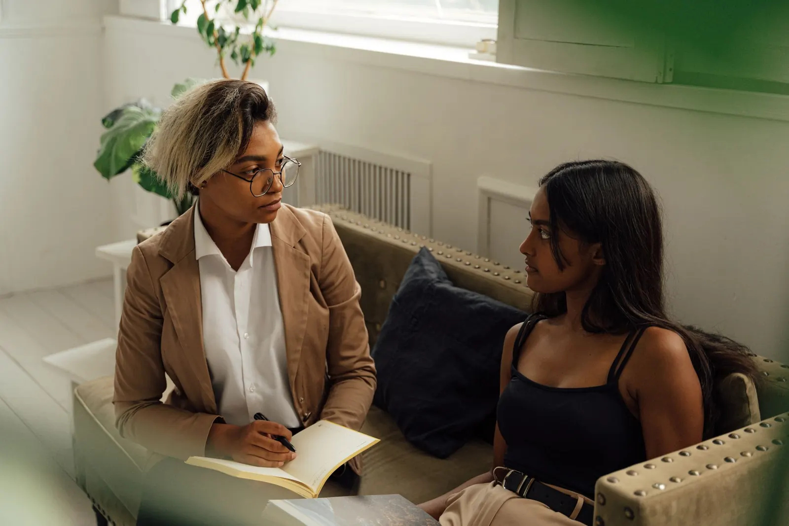 Deux femmes en conversation dans un salon lumineux, l'une prend des notes dans un carnet