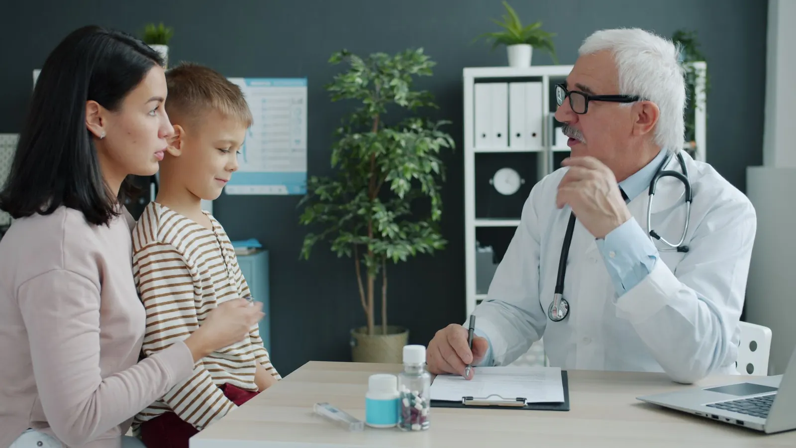 Médecin âgé consultant une mère et son jeune fils dans un cabinet, un dossier posé sur le bureau