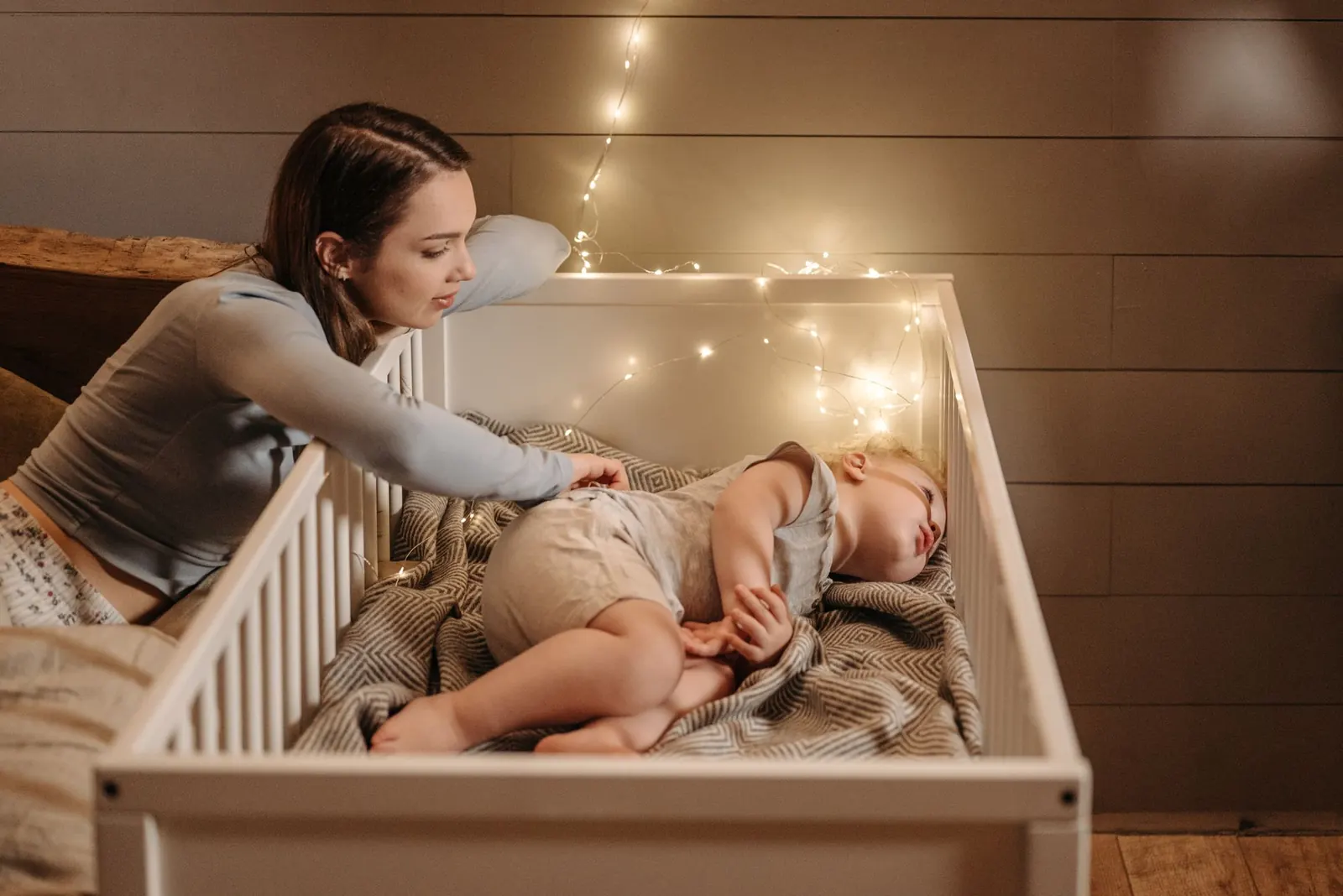 Une jeune mère penchée sur le lit à barreaux de son bébé endormi, dans une chambre éclairée par une guirlande lumineuse