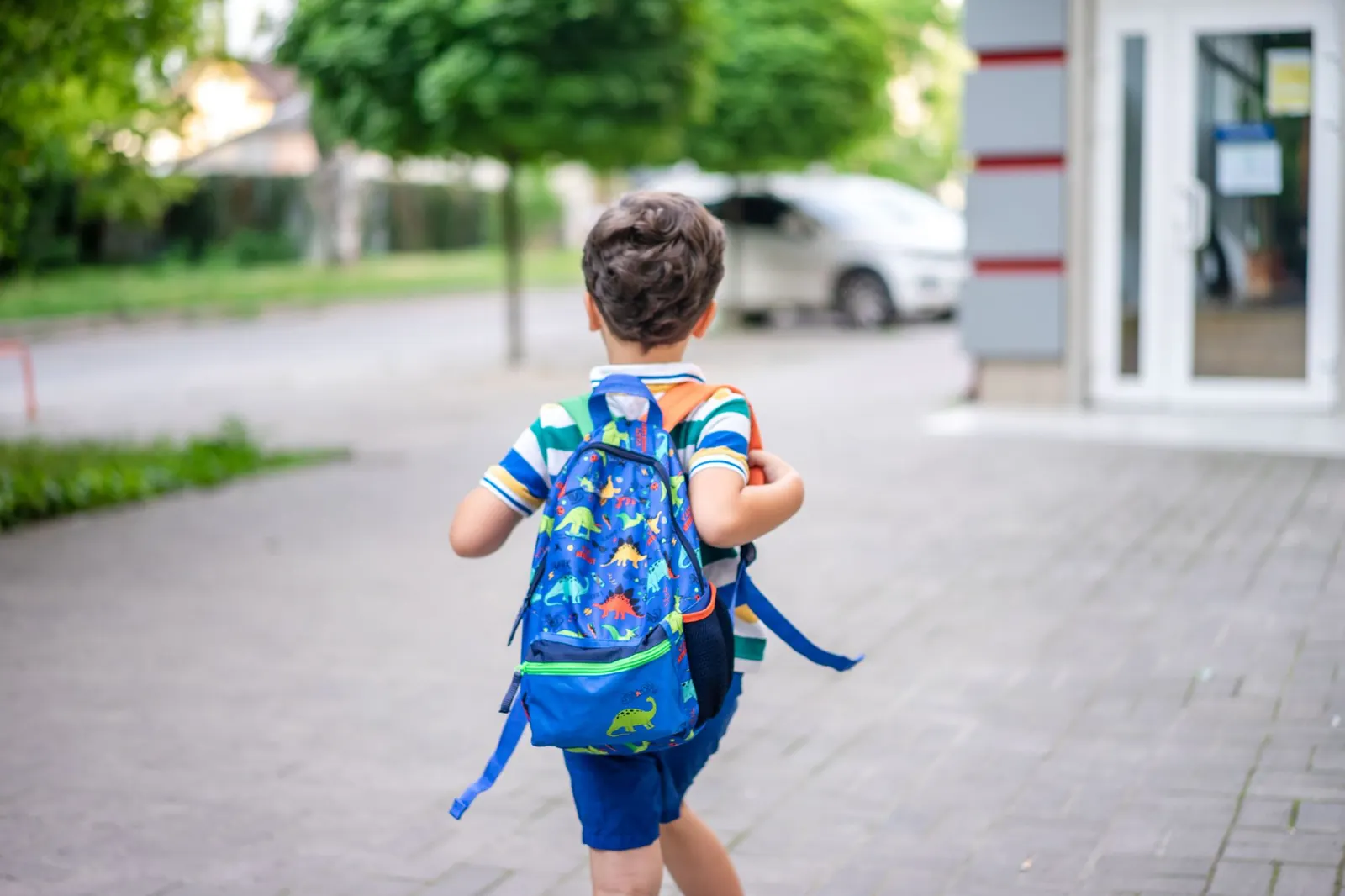 Petit garçon de dos avec un cartable bleu à motifs de dinosaures, marchant sur un trottoir devant une école