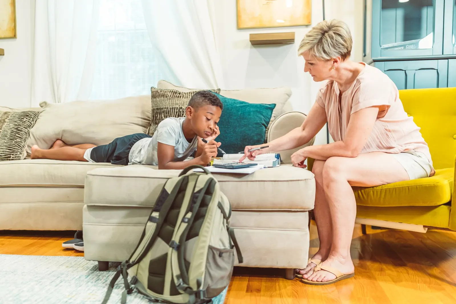 Une mère aide son fils à faire ses devoirs dans le salon, un sac à dos posé devant le canapé