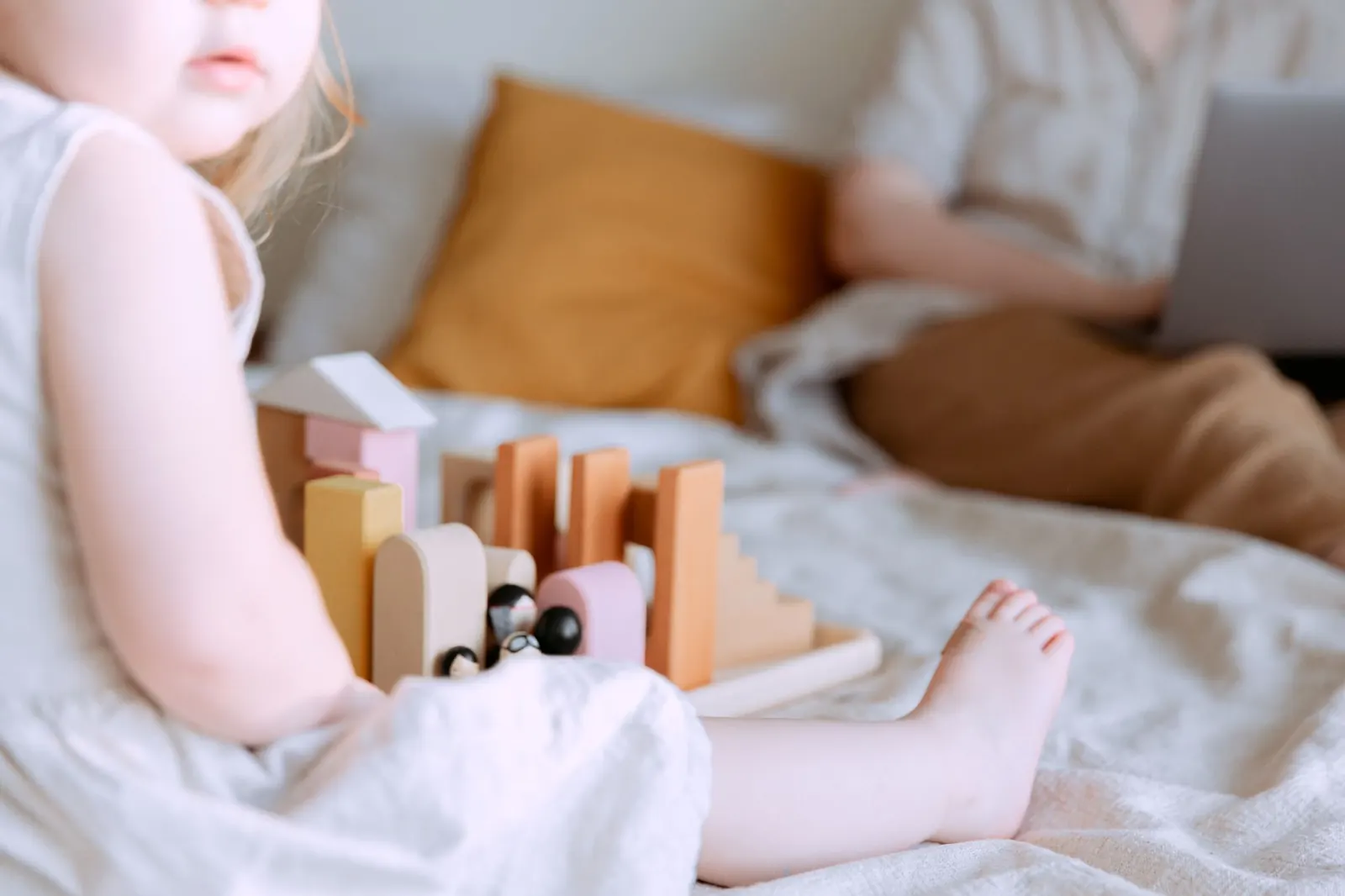 Une petite fille joue avec des cubes en bois sur un lit pendant qu'un parent travaille sur un ordinateur portable en arrière-plan
