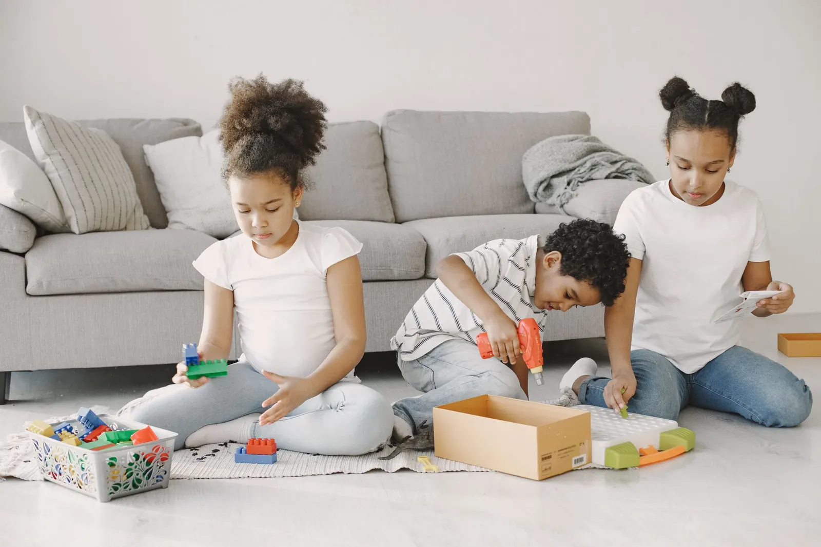 Trois enfants jouent avec des jouets éducatifs, assis sur le sol d'un salon devant un canapé gris