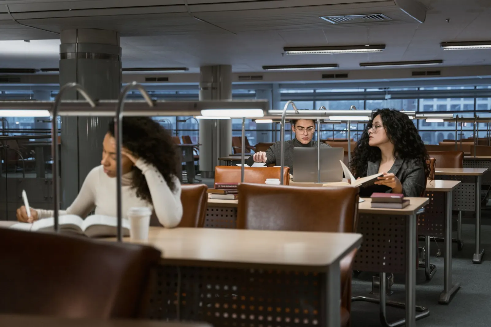 Étudiants installés à des tables de bibliothèque universitaire, livres et ordinateurs ouverts