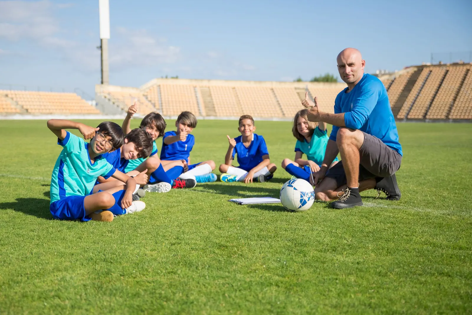 Entraîneur et jeunes footballeurs en maillots bleus, assis sur la pelouse d'un stade, le pouce levé