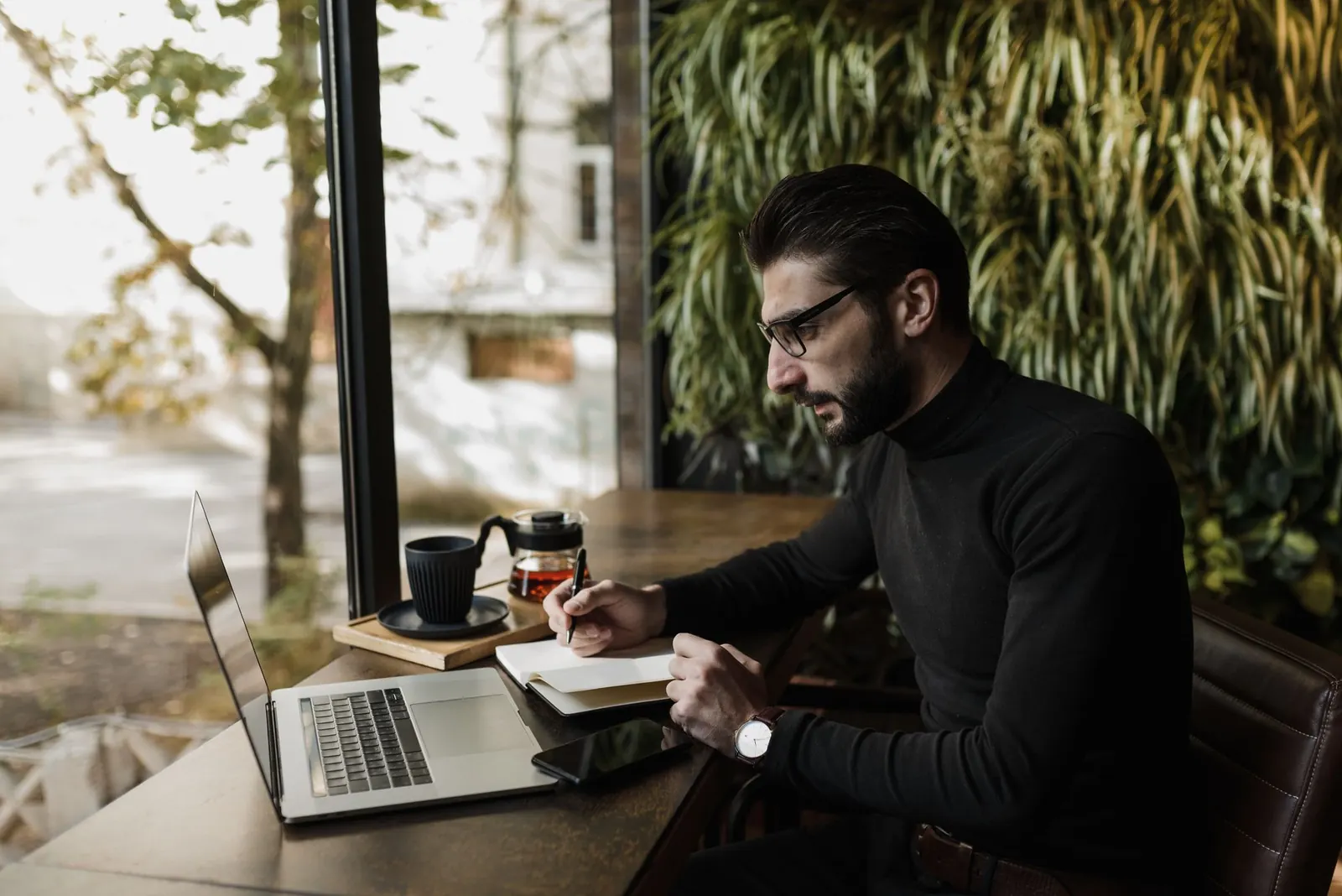 Homme assis dans un café devant un ordinateur portable, prenant des notes dans un carnet
